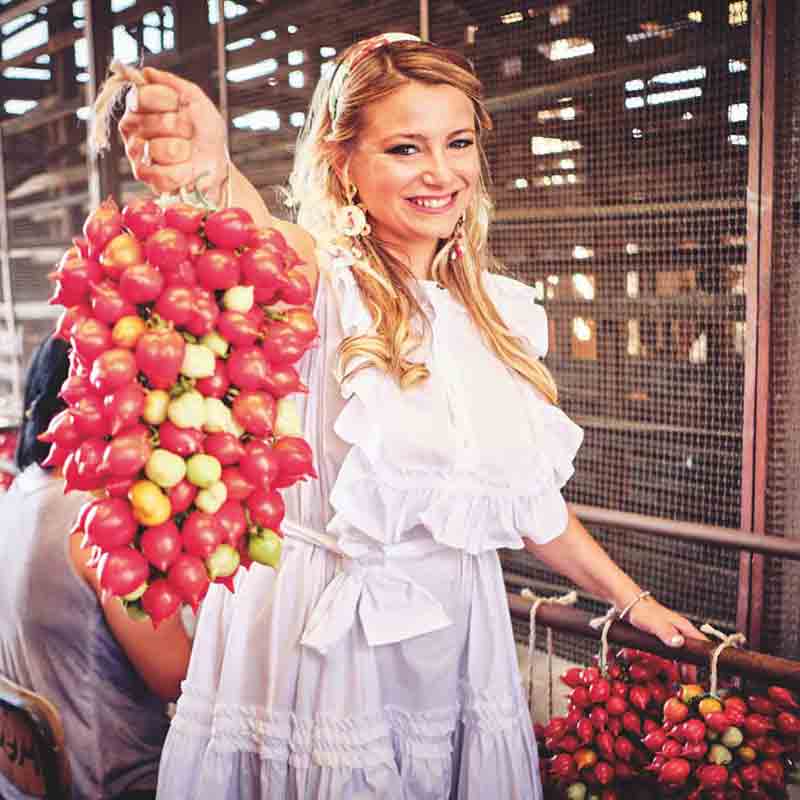 Diana, Inhaberin von Italianavera, trägt ein weißes Kleid und hält sonnenverwöhnte Tomaten in die Kamera.