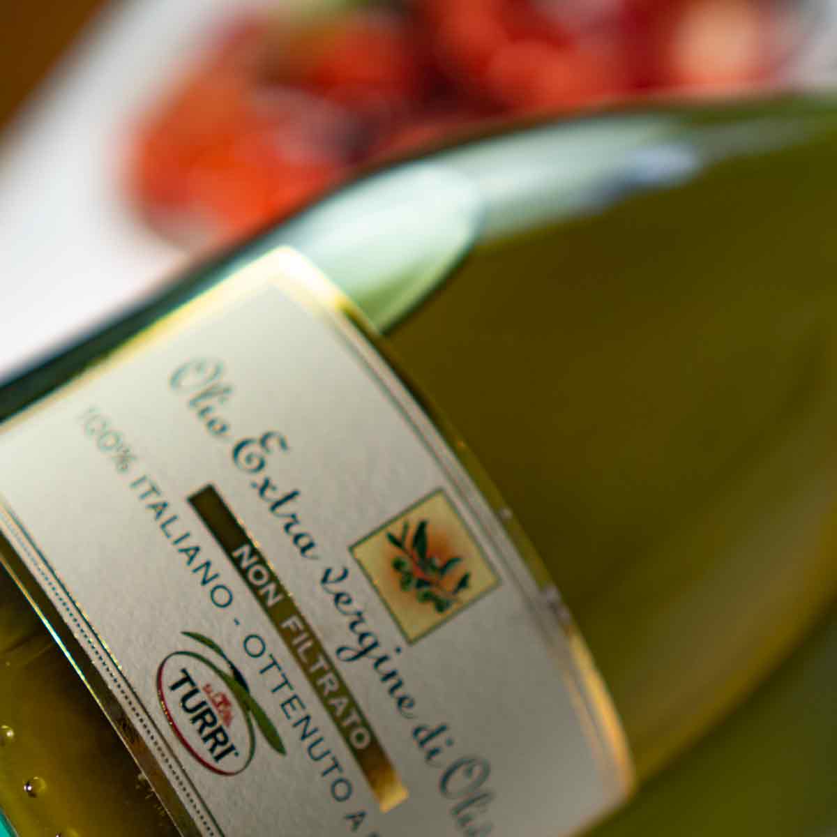 Turri Olivenölflasche vor unscharfem Hintergrund mit Tomaten.